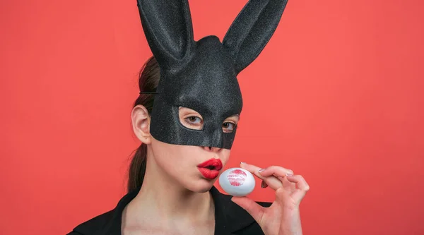 Sexy woman wearing a black Easter Bunny. Female mouth kiss. Red lip imprint on easter egg on red background.