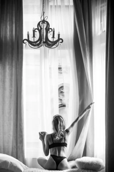 Vista posteriore di giovani donne in forma sana in camera da letto dopo il risveglio. Mattina a bere caffè. Donna che apre le tende delle finestre. Vista posteriore a donna sexy in appartamento di lusso o in camera d'albergo. — Foto Stock