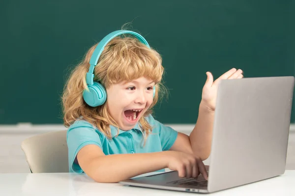 Opgewonden jongen in hoofdtelefoon zitten aan het bureau, online studeren op laptop op school, klever verbaasd kind dragen oortelefoons in notebook leren met behulp van internetlessen. — Stockfoto