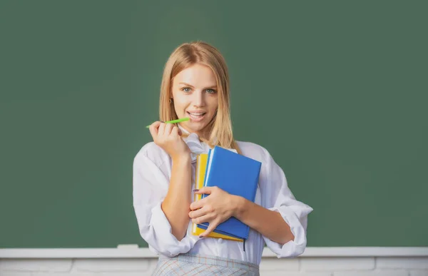 Egy fiatal főiskolás lány portréja, aki az osztályteremben tanul, táblás háttérrel. Kreatív fiatal mosolygós női diák tollal és könyvvel. — Stock Fotó