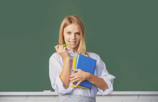 Retrato de una joven estudiante universitaria que estudia en clase con antecedentes en pizarra. Joven y creativa estudiante sonriente con pluma y libro. —  Fotos de Stock