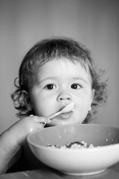 Funny baby eating food himself with a spoon on kitchen. Child nutrition concept. Funny child face closeup. — Stock Photo, Image