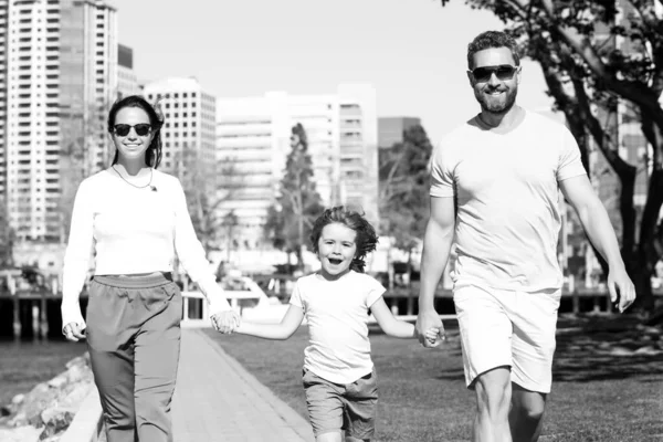 幸せなアメリカの両親は息子と手を取り合い、日当たりの良い夏の街の通り、柔らかい家族の瞬間を歩いています。家族旅行. — ストック写真