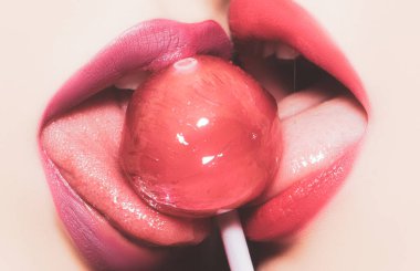 Şehvetli kadın ağzı yalıyor. Lezbiyen oral zevki. Yakından bakınca, ağzında pembe ve mor ruj izi olan ve yuvarlak kırmızı lolipop şekeri yalayan iki seksi, şehvetli kadın dudakları görünüyor..