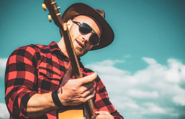 Homem com guitarra. Bonito jovem vestindo óculos de sol e tocando guitarra no fundo do céu. Homens vaqueiros. Camping ocidental. — Fotografia de Stock