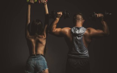 Kaba bir çift halterleriyle birlikte egzersiz yapıyor. Seksi, formda bir vücut. Çıplak vücutlu kaslı bir adam, arka planda koyu renkli halterleri olan fitness kadını. Dambılı çift eğitimi.