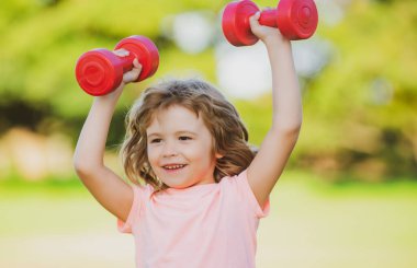 Küçük mutlu çocuk yeşil parkta dambıllarla egzersiz yapıyor. Dambılı, gülümseyen bir çocuğun yakın plan portresi. Mutlu çocuk dışarıda egzersiz yapıyor..