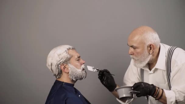 Hair coloring man. Making of a dyed hair for a bearded hipster guy. Beard coloring man agains grey hair. Bearded man, barber shop. Professional hairdresser drying hair in studio. — Stock Video