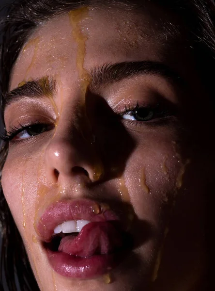Sensual young woman face with honey drop close up. Beautiful girl tongue licking honey lips. Sexy model with honey drip on face.