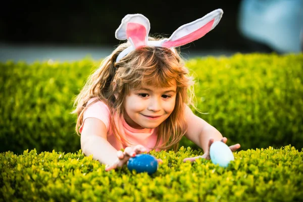 Niño cazando huevos de Pascua. Niño con traje de conejo con orejas de conejo cazando huevos de Pascua en la hierba en el parque de primavera. —  Fotos de Stock