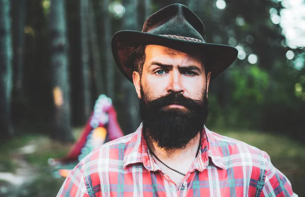 Homem maduro de camisa com barba de chapéu. Homem barbudo relaxa na floresta. Um homem barbudo sério. Atraente turista na floresta. Cuidado com a barba. Acampamento. Cowboy brutal com barba comprida. Cuidado barba de cabelo. — Fotografia de Stock