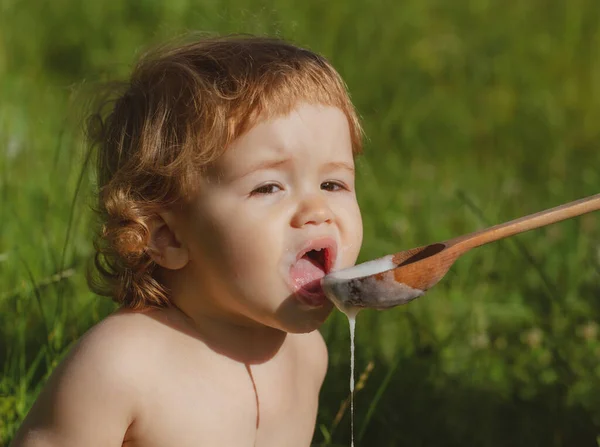 Funny child face. Feeding baby with big spoon outdoor on green grass. — Stock Photo, Image