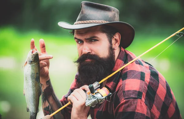 Pescatore serio vicino all'acqua con il pesce. Pescatore con canna da pesca. Attrezzatura da pesca dei pescatori. La pesca è divertente. Rilassati sulla natura. Pesci volanti hobby dell'uomo. Uomo barbuto . — Foto Stock