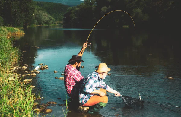 男性趣味の釣り。湖の朝の光の間にネットとロッドと一緒に釣りシャツに身を包んだ2人の男性の友人。川沿いの男は釣りをしながらのどかな風景を楽しむ. — ストック写真