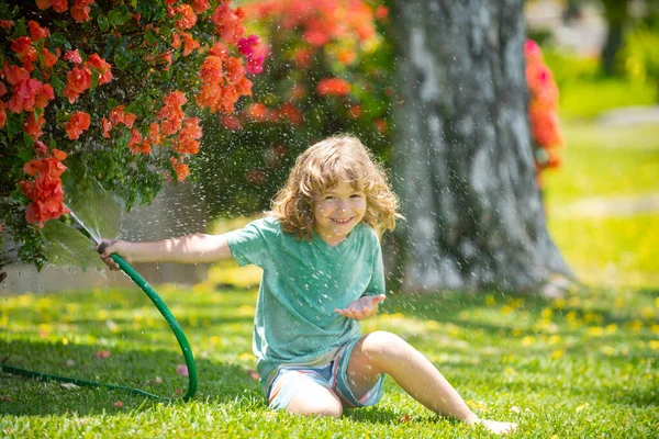 Милый мальчик поливает цветы в саду в летний день. Детский фермер с садовым шлангом в посадочном растении. Шланг. Забавный ребенок поливает растения во дворе сада. — стоковое фото