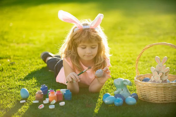 Пасхальный мальчик в кроличьих ушах охотится на пасхальные яйца на открытом воздухе. Пасхальные дети с кроличьими ушами и на фоне травы. — стоковое фото