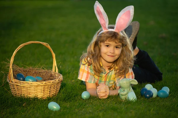 Niño con huevos de Pascua en cesta al aire libre. Niño tendido en el césped en el parque. Caza de huevos de Pascua. Cara de niños Fynny. Bunny niños con conejo orejas de conejo. —  Fotos de Stock