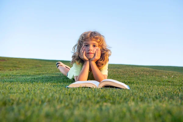 Kid läsa bok om gräs på gräs och himmel bakgrund med kopia utrymme. Rolig pojke läser boken i vårparken. — Stockfoto