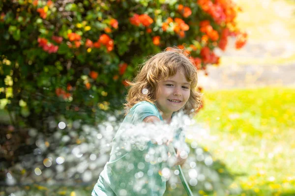 Το παιδί που παίζει στον κήπο, χύνει από το λάστιχο και βρέχει. Χαρούμενη παιδική ιδέα. Το παιδί ρίχνει νερό στα δέντρα. Παιδική φροντίδα για τα φυτά στην πίσω αυλή. — Φωτογραφία Αρχείου