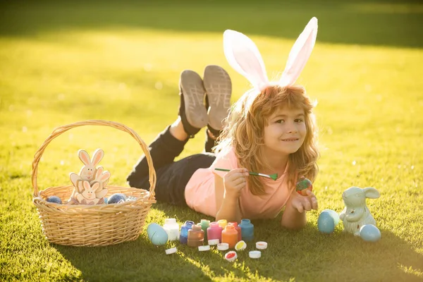 Мальчик в костюме кролика с кроличьими ушами рисует пасхальные яйца на траве в весеннем парке. — стоковое фото