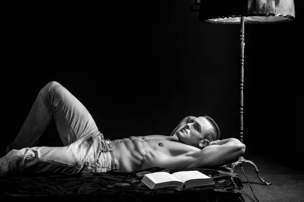 Pohledný muž v modrém džínovém s knihou a holým trupem. Ležel na posteli a odpočíval. Luxusní životní styl. — Stock fotografie