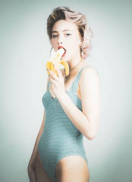 Niña pinup en traje de baño comiendo plátano, aislado en gris. Jovencita divertida comiendo plátano. Frutas tropicales. Concepto de verano. Alimentación saludable . —  Fotos de Stock