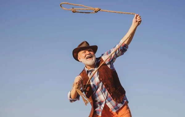 Old wild west cowboy at rodeo. Western pensioner with lasso rope. Bearded man with brown jacket and hat catching horse or cow. — Stock Photo, Image