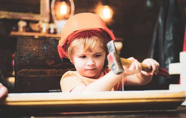 Kid hammers nails with a hammer in a wooden board. Boy child busy in helmet learning to use hammer. Child hammering nail into wood at home. — Stock Photo, Image