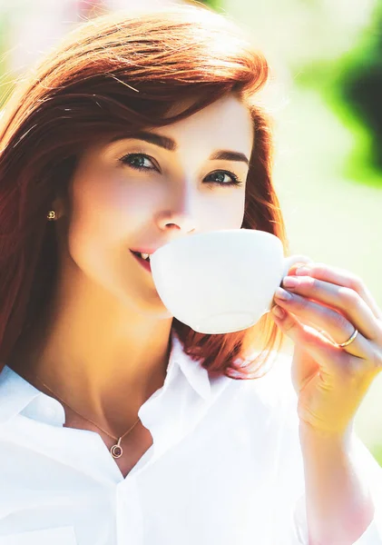 Ritratto ravvicinato di una donna che tiene una tazza di caffè calda all'aperto sullo sfondo della primavera. Testa rossa ragazza bere caffè . — Foto Stock