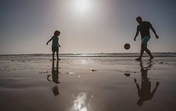 父と息子はビーチでサッカーやサッカーをしたり、日没のシルエットをしたりします。父と子供は屋外で楽しんでいます. — ストック写真