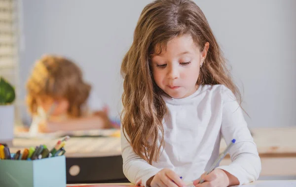Vicces tanuló az iskolában. Egy kislány ül az asztalnál az osztályban. Egy fiatal iskolás lány portréja, aki jegyzetfüzetben tanul és ír. Koncentrált kisiskolás gyerekek szórakoztató, írás. — Stock Fotó
