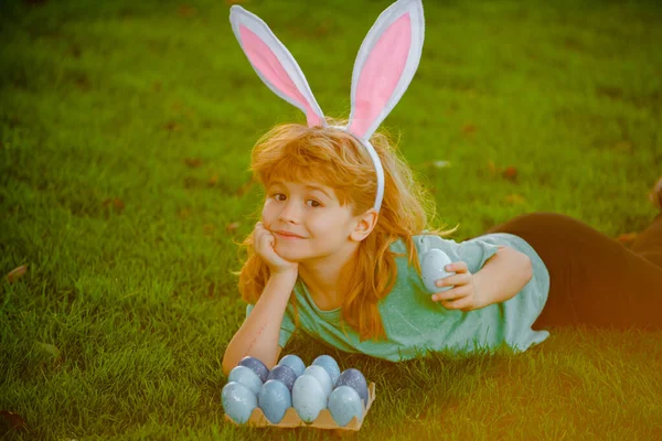 Niño cazando huevos de Pascua. Niño en orejas de conejo recogiendo huevos de Pascua, poniendo en la hierba. — Foto de Stock