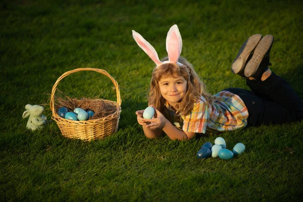 Conejito con orejas de conejo. Niños cazando huevos de Pascua. Niño acostado en la hierba y encontrando huevos de Pascua. Niño con huevos de Pascua y orejas de conejo, retrato al aire libre. —  Fotos de Stock