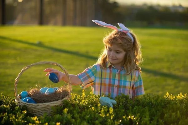 Mały króliczek z króliczymi uszami. Chłopiec z wielkanocnymi jajkami w koszyku na świeżym powietrzu. Polowanie na pisanki. Fynny dzieci twarz. — Zdjęcie stockowe