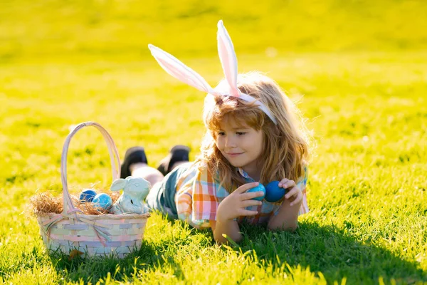 Niño con traje de conejo con orejas de conejo cazando huevos de Pascua que ponen en la hierba en el parque. — Foto de Stock