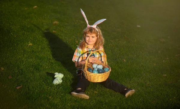 Παιδί με πασχαλινά αυγά στο καλάθι υπαίθρια. Αγόρι στο γρασίδι στο πάρκο. Κυνήγι πασχαλινών αυγών. Το πορτραίτο της Φίνι. Χαριτωμένο παιδί λαγουδάκι με αυτιά κουνέλι. — Φωτογραφία Αρχείου