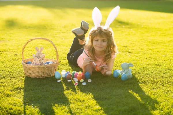 Niño con huevos de Pascua y orejas de conejo pintando huevos al aire libre. Lindo niño teniendo feliz Pascua en el parque. — Foto de Stock