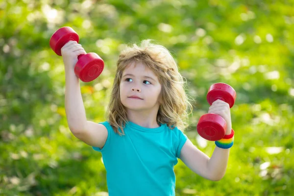 Junge, der eine Hantel hochhält. Niedliches Kindertraining mit Kurzhanteln. Kinderfitness. Junge turnt mit Kurzhanteln im Freien. Lustig aufgeregter Kindersportler. — Stockfoto