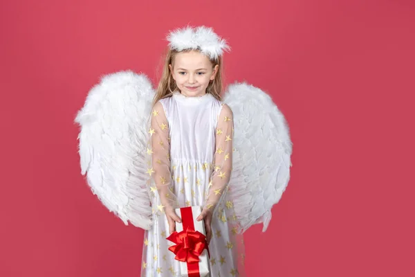 現在の贈り物、スタジオの肖像と子供の女の子の天使。白い翼を持つ小さな天使は贈り物を保持します。天使の翼を持つかわいい天使の子の女の子,赤に隔離されました.バレンタインギフトカード. — ストック写真