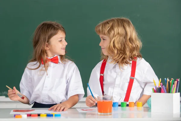 Roliga elever ritar i klassrummet på skolans svarta tavlan bakgrund. Skolkamrater barn pojke och flicka målning tillsammans i klassen. Bästa barn vänner njuter av vänskap. — Stockfoto