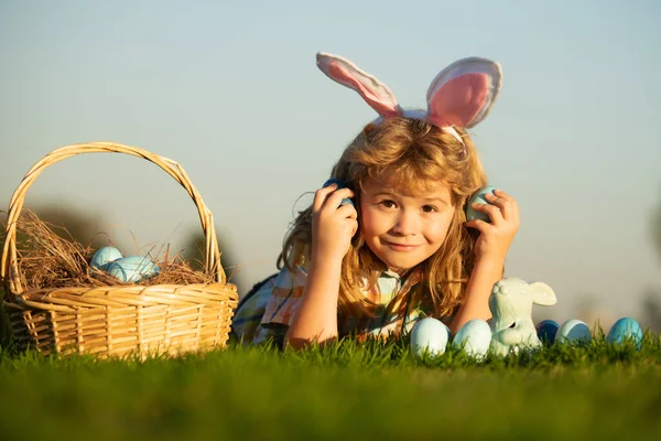 Niños niño cazando huevos de Pascua en el parque que pone en la hierba en el fondo del cielo con espacio de copia. Niño con orejas de conejo. —  Fotos de Stock