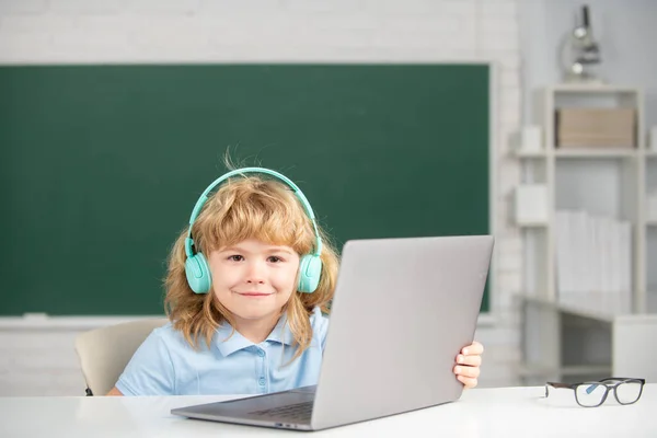 Bonito menino sorrindo usando fones de ouvido, estudar com laptop em sala de aula, ouvir curso de aula de áudio. Ensino fundamental e educação. — Fotografia de Stock