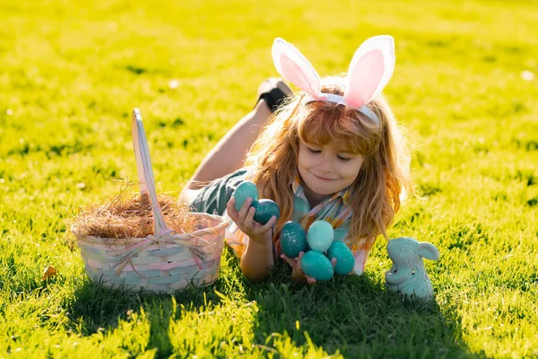 Barnpojke jagar påskägg, lägger på gräs. Grabben i kaninöron utomhus. Påskhare. Vårlovet. — Stockfoto