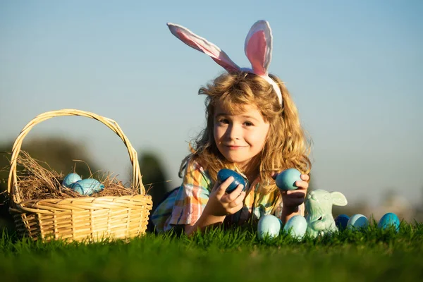 Мальчик с пасхальными яйцами и корзиной на траве. Дети охотятся на пасхальные яйца, откладывают на траву. Дети пасхальных кроликов на фоне неба с копировальным пространством. — стоковое фото