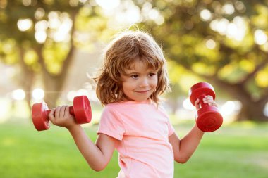 Yeşil parkta dambıllarla egzersiz yapan sevimli küçük çocuk. Dambılı spor bir çocuğun yakın plan portresi. Mutlu çocuk dışarıda egzersiz yapıyor..