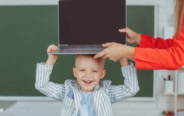 Szczęśliwy uśmiechnięty chłopiec trzyma laptopa na głowie podczas lekcji online w klasie, edukacja online. — Zdjęcie stockowe