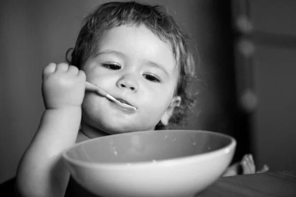 プレートのスプーンを閉じ保持から食べる面白い小さな赤ちゃんの少年の肖像画。口の中にスプーンを押しながら食べる赤ん坊. — ストック写真