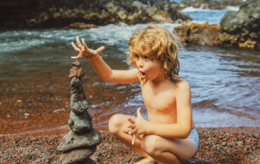 Komik çocuk kaplıca, denge, meditasyon ve zen teması için deniz zeminine karşı çakıl taşı yığını yapıyor. Çocuk sahilde oynuyor..