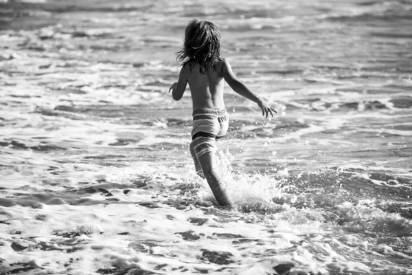 Πίσω όψη του παιδιού αγόρι έχουν τη διασκέδαση στην τροπική παραλία της θάλασσας. Αστείο παιδί τρέχει με πιτσιλιές από νερό στην άκρη του σέρφινγκ. Παιδική δραστηριότητα στις καλοκαιρινές διακοπές. — Φωτογραφία Αρχείου