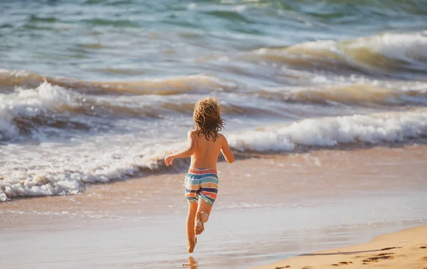 Ragazzo felice divertirsi sulla spiaggia tropicale del mare. Bambino divertente correre con spruzzi di acqua piscina lungo il bordo surf. Attività per bambini durante le vacanze estive. — Foto Stock